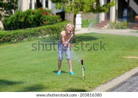 Happy laughing teenage boy in stripes swimming shorts running though garden sprinkler playing with water splashes having fun in the lawn on a sunny hot summer vacation day