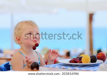 Happy healthy little child, cute blonde toddler girl, enjoying summer vacations eating sweet cherries and other fruits sitting in high chair at beach restaurant with sea view in tropical resort