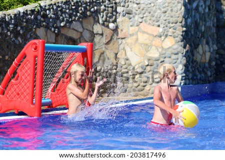 Two happy teenage boys, sportive twin brothers, having fun together playing waterpolo with colorful inflatable ball in outdoors swimming pool in aquapark during summer sea vacation in tropical resort