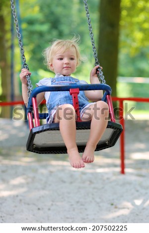 Happy little child, smiling blonde toddler girl in casual outfit having fun on a swing enjoying a warm sunny summer day on a playground in a park