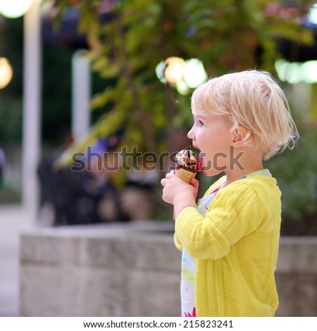 Happy little child, adorable blonde toddler girl in beautiful dress, walking on crowded touristic street in the center of the city eating ice-cream on a sunny summer evening