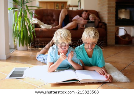 Happy family at home, bright sunny classic living room with tiles floor and big windows - two brothers reading lying on the floor, father with daughter playing on sofa at background