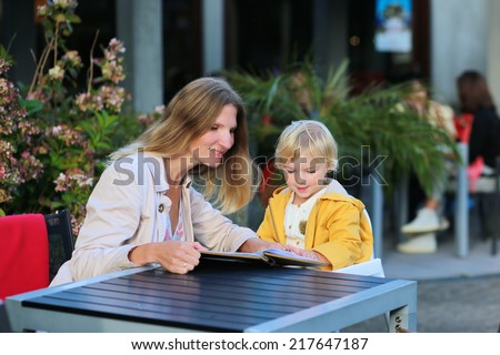 Happy family of two, mother, attractive young woman and her daughter, adorable toddler girl enjoying lunch at a beautiful outside cafe choosing meal from menu card on a sunny early autumn day