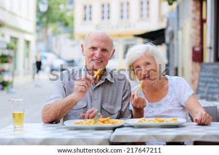 Happy active senior couple enjoying time together eating belgian french fries in outdoors street cafe on a summer day in typical European town - active retirement concept