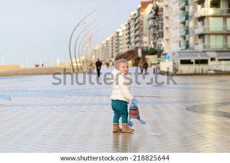 Adorable little toddler girl in warm white coat and colorful hat walks on the promenade along the beach on a sunny early spring or late autumn day at sunset, North Sea, Belgium, De Panne