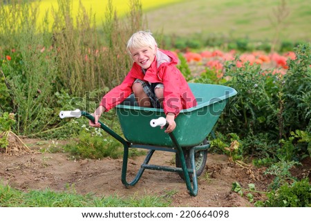 Happy child, laughing school boy in red raincoat and rubber boots, enjoying nature sitting in wheelbarrow helping to harvest bio pumpkins growing in organic field on sunny autumn day