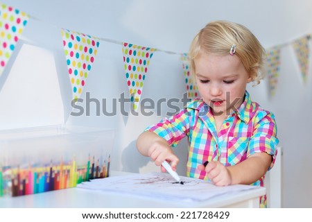 Active little preschool age child, cute toddler girl with blonde curly hair, drawing picture on paper using colorful pencils and felt-tip pens, sitting at white table indoors at home or kindergarten