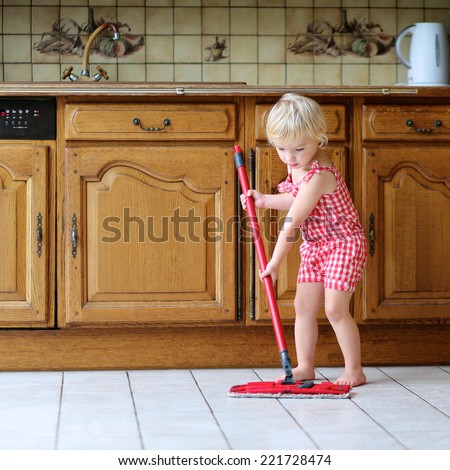Cute blonde toddler girl helping with housekeeping in classical kitchen with wooden cabinetry cleaning tiles floor with wet flat mop