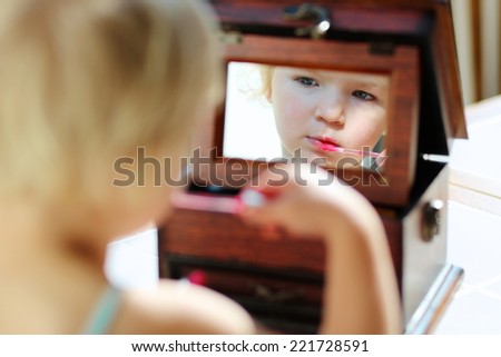 Little child, beautiful toddler girl with blonde curly hair, applying mom\'s pink lipstick to her lips looking at the mirror