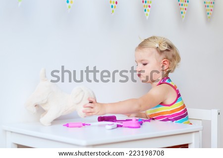 Little child, adorable blonde toddler girl, playing doctor role game and treating her puppy using different medical tools sitting at small white table in playroom at home, school or kindergarten