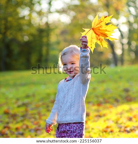 Happy little child, cute toddler girl with curly blonde hair wearing yellow jacket playing with colorful leaves in park on sunny warm autumn day