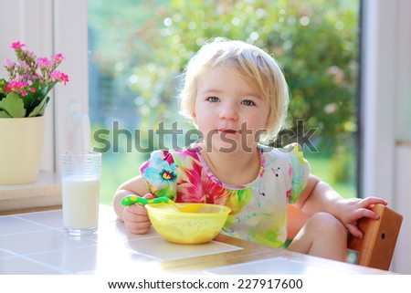 Happy little child, blonde curly toddler girl, enjoying healthy breakfast eating oatmeal porridge and drinking milk sitting in high chair at bright sunny kitchen next to big garden view window