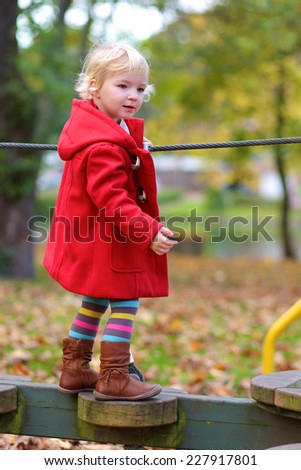 Happy active little child, blonde curly toddler girl wearing beautiful red duffle coat, having fun at playground in the park on sunny autumn day