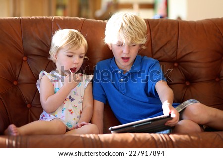 Two happy blonde kids, brother and sister, teenage boy and cute toddler girl, playing together with tablet pc sitting indoors at home in sunny living room on brown leather sofa
