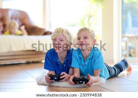 Group of kids twin teenage brothers having fun after school day playing video game holding joysticks in hands lying cozy on tiles floor on warm lambskin in bright sunny living room with big windows