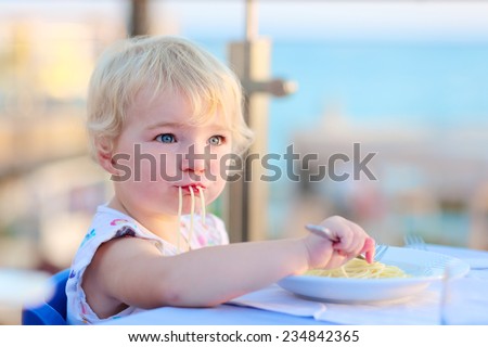 Happy healthy little child, cute blonde toddler girl, enjoying summer vacations in tropical resort eating pasta at beach restaurant with sea view