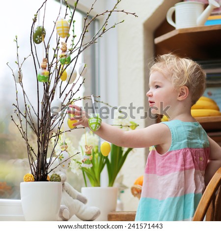 Adorable child, little blonde toddler girl in beautiful dress decorating with Easter eggs cherry tree branches standing in the kitchen next a window with garden view