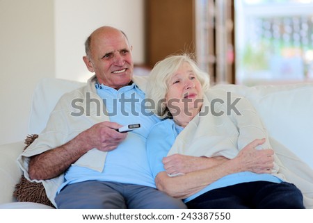 Happy senior couple relaxing at home sitting on sofa wrapped in soft plaid and watching tv
