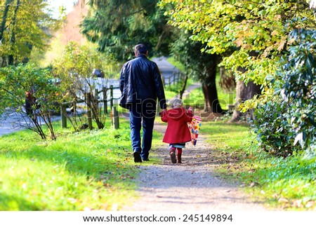 Rear view of active father and little child, blonde toddler girl in beautiful red coat with colorful umbrella walking hand in hand in autumn or spring park on a sunny day