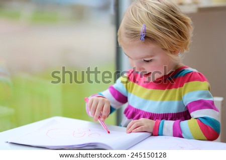 Cute little child, blonde preschooler girl is drawing with felt-tip pens at home or kindergarten sitting at small table next to window