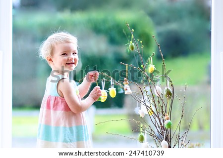Cheerful little child, blonde curly toddler girl decorating cherry tree branches with Easter eggs standing indoors next to a big window with street view