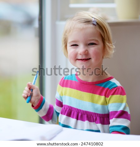 Portrait of cute little child, blonde preschooler girl holding felt-tip pen drawing on paper sitting at small table next to big window at home or kindergarten