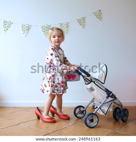 Happy little child, cute blonde toddler girl, wearing beautiful dress and red mom\'s shoes playing role game pushing stroller with baby doll indoors at home or kindergarten
