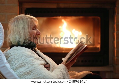 Happy senior woman, wrapped in warm knitted plaid, relaxing at home in the evening, sitting in rocking chair by fireplace and reading a book - successful retirement concept