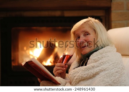 Happy senior woman, wrapped in warm knitted plaid, relaxing at home in the evening, sitting in rocking chair, drinking wine, reading book and enjoying fireplace - successful retirement concept