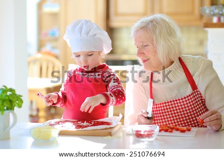 Happy family, grandmother with granddaughter, adorable little girl, preparing delicious pizza topping it with tomato sauce, vegetables and cheese, sitting at white dining table at bright sunny room