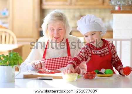 Happy family, grandmother with granddaughter, adorable little girl, preparing delicious pizza topping it with tomato sauce, vegetables and cheese, sitting at white dining table at bright sunny room