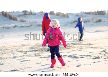 Active little child, laughing cute toddler girl wearing warm pink coat and pretty shoes playing with her brothers on the beach of North Sea on sunny winter or spring day