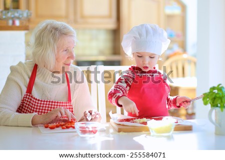 Happy family, grandmother and granddaughter, adorable little girl, preparing delicious pizza together topping it with tomato sauce, vegetables and cheese, sitting at table at bright sunny room at home