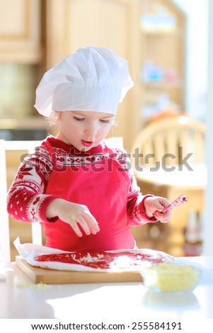 Smiling little child, adorable toddler girl wearing red chef apron and white hat, preparing delicious pizza for family party topping it with tomato sauce, vegetables and cheese, sitting at sunny room