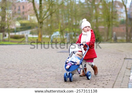 Stylish little child, cute blonde toddler girl, wearing beautiful red coat playing role game pushing stroller with toy outdoors in the city park