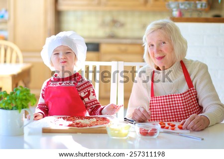 Happy family, grandmother with her granddaughter, adorable little girl, preparing delicious pizza together topping it with tomato sauce, vegetables and cheese. Selective focus on child