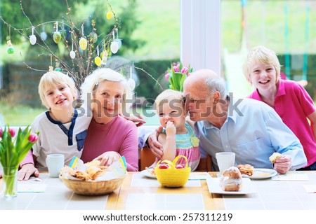 Family of 5: grandparents and grandchildren, teenage boys and toddler girl eating eggs and pastry enjoying family breakfast on Easter day sitting together in sunny kitchen with big garden view window