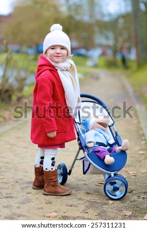 Stylish little child, cute blonde toddler girl, wearing beautiful red coat playing role game pushing stroller with toy outdoors in the city park