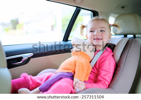 Portrait of happy little child, cute blonde toddler girl sitting comfortable in car seat with safety belts enjoying ride in the auto on family weekend trip
