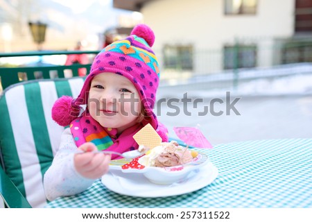 Lovely little child, adorable toddler girl wearing colorful pink hat, eating delicious ice cream in outdoors street cafe enjoying weekend city trip with family