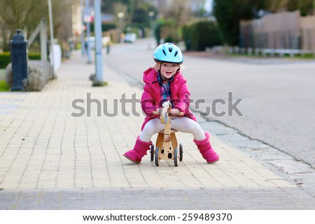 Happy little kid, cute blonde toddler girl in pink jacket and blue safety helmet playing outdoors on the street riding her push bike, wooden horse with three wheels