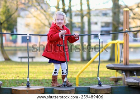 Happy active little child, blonde curly toddler girl wearing beautiful red coat, having fun playing at playground in city park on sunny spring day