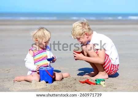 Two happy active children, teenage boy with his little sister, cute blonde toddler girl, playing with plastic toys building sand castles sitting on wide sandy North European beach