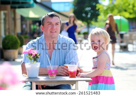 Happy family of two, smiling man with his cute toddler daughter, relaxing together in summer outdoors cafe drinking coffee, juice or smoothie and eating muffin - father and child, parenting concept