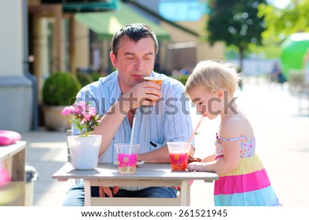 Happy family of two, smiling man with his cute toddler daughter, relaxing together in summer outdoors cafe drinking coffee, juice or smoothie and eating muffin - father and child, parenting concept