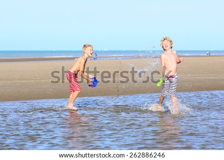 Two happy laughing kids, teenage boys, twin brothers having fun at the North Sea playing together on the beach, running and splashing water from bucket and plastic guns