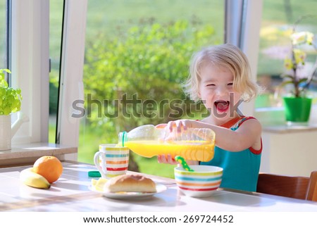 Happy child of preschool age, blonde curly toddler girl, enjoying healthy breakfast eating sandwich and fruits and drinking orange juice sitting at bright sunny kitchen next to big garden view window