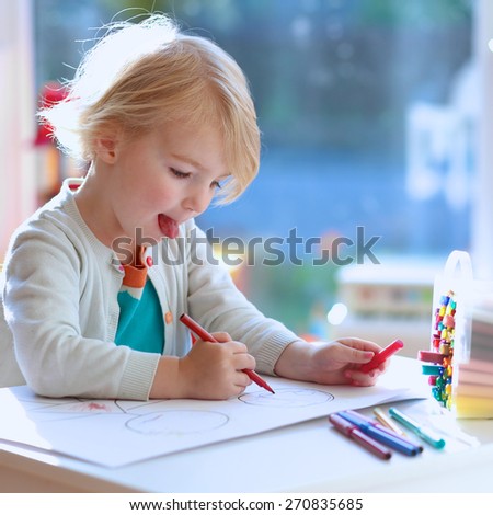 Cute little child, blonde preschooler girl is drawing and painting with colorful felt-tip pens at home or kindergarten sitting at small table in bright sunny playroom