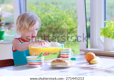 Happy child of preschool age, blonde curly toddler girl enjoying healthy breakfast eating sandwich and fruits and drinking orange juice sitting at bright sunny kitchen next to big garden view window