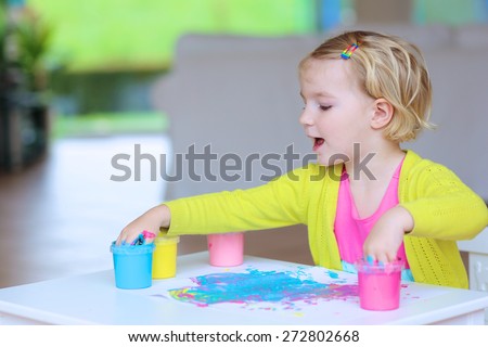 Cute little child, blonde artistic toddler girl painting with colorful finger paints indoors at bright room at home or kindergarten